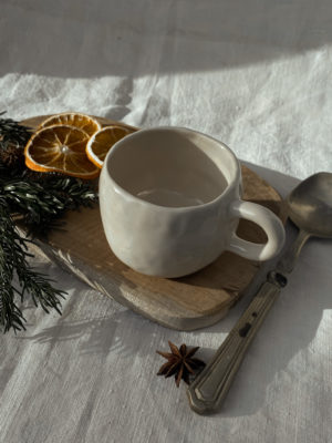 tasse crème vu du haut de 3/4 sur un rondin de bois avec oranges séchées et branches de sapin sur le coté + cuillère.